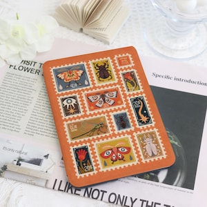 Puede incluir: Un cuaderno naranja con diseños de sellos postales que ilustran insectos y flores. Los sellos tienen diferentes valores y bordes decorativos. El cuaderno está sobre una superficie blanca.