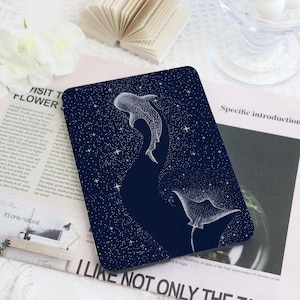 Puede incluir: Un posavasos rectangular azul oscuro con una ilustración blanca de un tiburón ballena y una manta raya en un cielo estrellado. El posavasos tiene esquinas redondeadas. Texto visible alrededor.