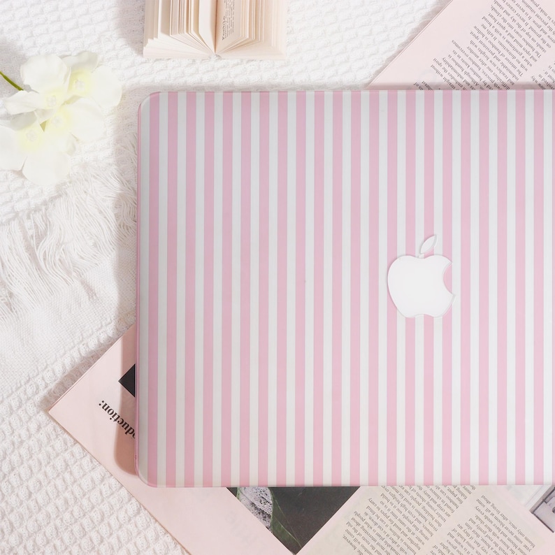 May include: A pink and white striped laptop with the Apple logo. The laptop is open and resting on a white surface. The background includes a book and a flower.