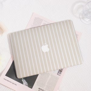 Puede incluir: Una funda para portátil con rayas beige y blancas y el logotipo de Apple. La funda está sobre un periódico rosa. El fondo es una superficie blanca texturizada.