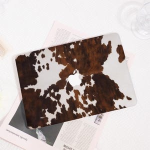 May include: A laptop skin featuring a brown and white cowhide print pattern. The design covers the exterior lid of the laptop, leaving a circular cutout in the centre that reveals the white Apple logo. The surface has a smooth, matte finish.