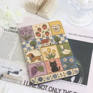 May include: A rectangular e-reader case featuring a grid of colorful illustrations including purple tulips, red strawberries, a teacup, a house, a dachshund, yellow tulips, a white duck, grapes, and a black cat, all bordered by floral patterns.