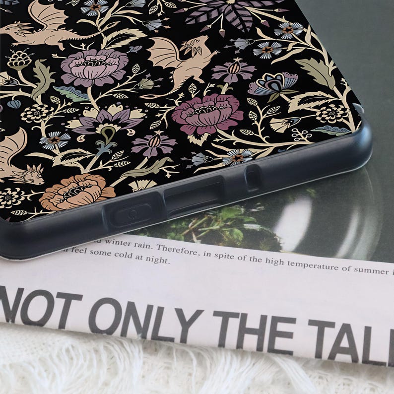 May include: Black phone case featuring a floral and dragon pattern in shades of beige, purple, and blue. The design includes stylized flowers and winged dragons. The case is on top of a newspaper with the text "NOT ONLY THE TALI".