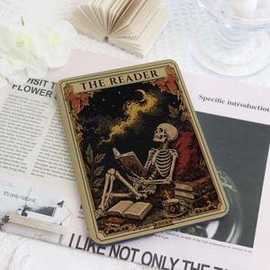 Könnte beinhalten: Eine rechteckige Metalldose mit einer Illustration im Tarot-Stil mit der Aufschrift THE READER. Das Bild zeigt ein Skelett, das an ein rotes Kissen gelehnt ein Buch unter einem Sternenhimmel mit Mondsichel liest, dazu Bücherstapel und eine Tasse.