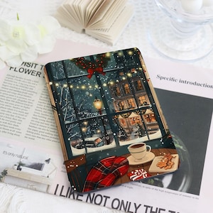 May include: A decorative notebook featuring a winter scene with a cozy cafe interior. The illustration shows a snowy night with a view of illuminated buildings, a cup of coffee, and gingerbread cookies. A red and black plaid blanket is also visible.
