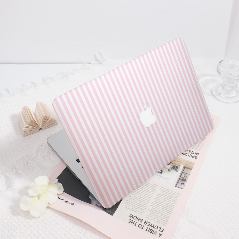 May include: A laptop with a pink and white striped cover, featuring the Apple logo. The laptop is open, resting on a magazine with the text "A VISIT TO THE FLOWER SHOW". A small paper book and white flowers are nearby.