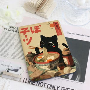 Può includere: Un taccuino rettangolare con un gatto nero che mangia ramen con le bacchette. Il design include testo giapponese, un sole e ciotole di cibo. Lo sfondo è beige con accenti marroni e rossi.