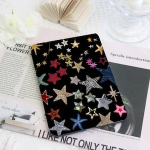 May include: Black rectangular case decorated with various star shapes in gold, silver, red, and other colors. Some stars have unique patterns, and some are glittery. The case is on a white surface.