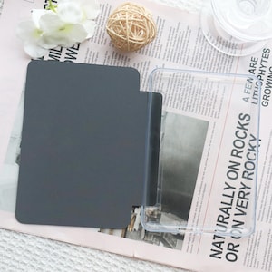 May include: A gray, rectangular notebook with rounded corners, next to a clear plastic case. The notebook is on a newspaper with the text "NATURALLY ON ROCKS OR ON VERY ROCKY". A decorative ball of twine and a white flower are also visible.