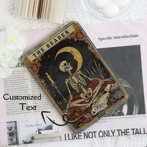 K&ouml;nnte beinhalten: Rechteckige Tarotkarte mit einem Skelett unter einem Mond und Sternen. Die Karte tr&auml;gt oben die Aufschrift "THE READER" und unten "Coral BOOKS". Das Design hat einen Vintage- und Gothic-Stil.