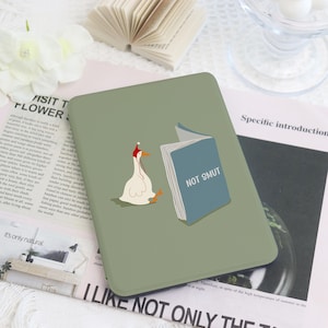May include: A sage green e-reader cover featuring a graphic of a white goose wearing a red hat, standing next to an open blue book with the text NOT SMUT printed on the cover. The design is centered on the rectangular protective case.