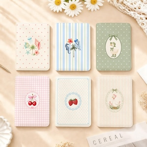 Puede incluir: Seis cuadernos rectangulares con esquinas redondeadas, con varios patrones que incluyen lunares, rayas y cuadros vichy. Las portadas muestran ilustraciones de vida marina, un pato con capota, cerezas, fresas y un patito en un marco floral.