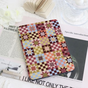 May include: A rectangular e-reader cover featuring a repeating quilt-inspired pattern of alternating star shapes and checkered squares in shades of maroon, mustard yellow, light blue, and soft peach, with various geometric textures within each block.