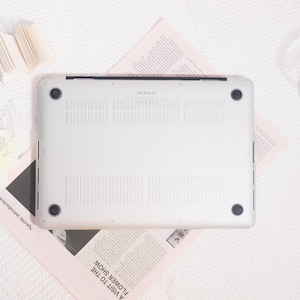 May include: Silver MacBook Pro laptop computer, viewed from the bottom, with ventilation slots and black rubber feet. The text "MacBook Pro" is visible. The laptop rests on a pink newspaper with the headline "A Visit to the Flower Show."