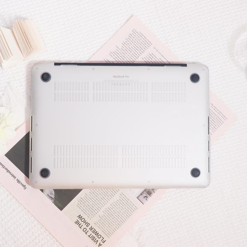 May include: Silver MacBook Pro laptop computer, viewed from the bottom, with ventilation slots and black rubber feet. The text "MacBook Pro" is visible. The laptop rests on a pink newspaper with the headline "A Visit to the Flower Show."