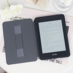 May include: A black Kindle e-reader with a gray cover, open to display text. The cover has a built-in stand. The Kindle is on a light pink surface with text and a white floral arrangement.