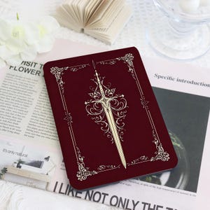 May include: A burgundy-colored notebook with an ornate sword design in cream. The cover features a decorative border and the text "I LIKE NOT ONLY THE" at the bottom. The notebook is on a patterned surface.
