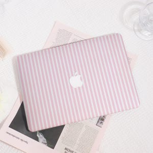 May include: A pink and white striped laptop case with the Apple logo. The case rests on light pink paper and a white textured surface. The case is designed to fit a laptop.