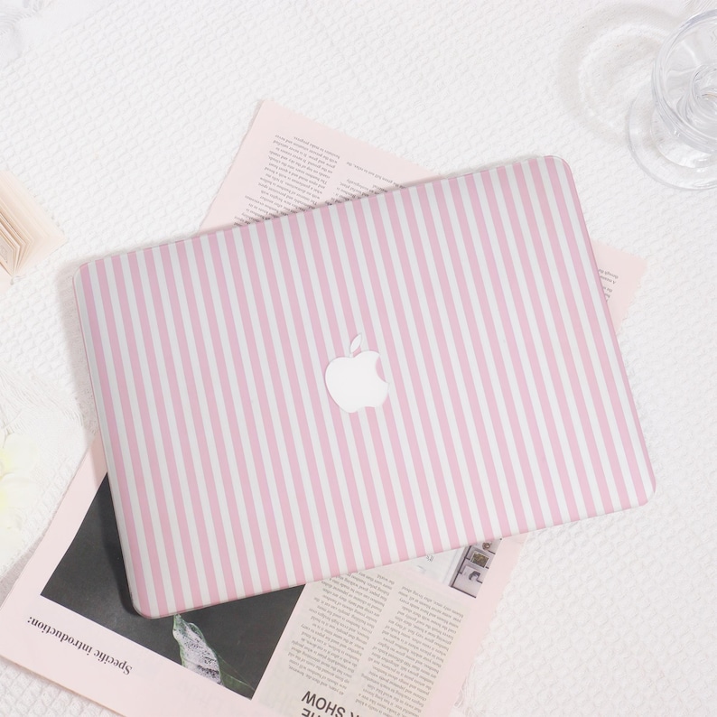 May include: A pink and white striped laptop case with the Apple logo. The case is on top of a light pink paper and a white textured surface. The case is designed to fit a laptop.