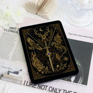 Pode incluir: Uma capa retangular preta para e-reader com uma ilustração dourada de uma espada central com uma gema azul, ladeada por um veado, um dragão, uma águia e uma rosa, tudo emoldurado por uma borda intrincada com uma lua crescente no topo.