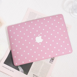 May include: A pink laptop case with a white polka dot pattern. The case features a white Apple logo in the center. The laptop case is resting on top of a newspaper.