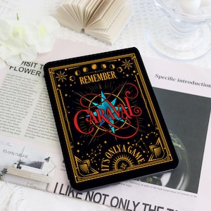 May include: A black rectangular card with gold trim and the words "REMEMBER CARAVAL" in red and blue. The card has a celestial design with stars and a compass rose. The text "IT'S ONLY A GAME" is also visible.