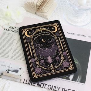 Puede incluir: Una funda para lector electrónico negra con un marco dorado ornamentado y motivos celestiales, incluyendo lunas y estrellas. La ilustración central muestra un paisaje montañoso nocturno con el texto, To the stars who listen, and the dreams that are answered.