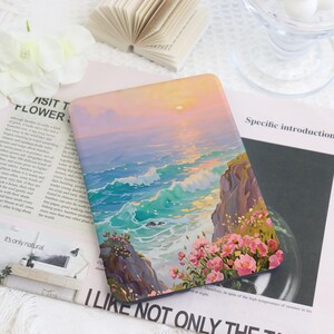 May include: A rectangular e-reader cover featuring a vibrant illustration of a coastal sunset. The artwork depicts turquoise ocean waves crashing against rocky cliffs adorned with pink flowers under a soft orange and purple sky.
