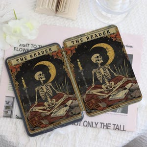 K&ouml;nnte beinhalten: Zwei Handyh&uuml;llen mit einem Tarotkarten-Design. Jede H&uuml;lle zeigt ein Skelett, einen Mond und Kerzen vor einem dunklen Hintergrund. Der Text "THE READER" steht oben. Die H&uuml;llen liegen auf einer hellrosa Oberfl&auml;che.