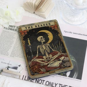 Può includere: Una carta dei tarocchi con uno scheletro seduto davanti a una luna crescente e stelle. La carta, intitolata "THE READER", ha uno sfondo scuro con una candela e libri. Il bordo della carta è un mix di oro e marrone.