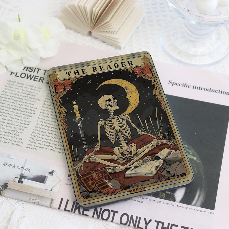 K&ouml;nnte beinhalten: Eine Tarotkarte mit einem Skelett, das vor einem Halbmond und Sternen sitzt. Die Karte mit dem Titel "THE READER" hat einen dunklen Hintergrund mit einer Kerze und B&uuml;chern. Der Rand der Karte ist eine Mischung aus Gold und Braun.