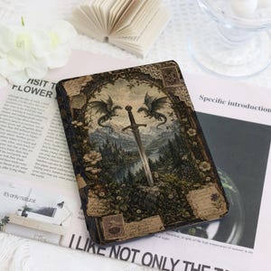 May include: A rectangular e-reader case featuring a detailed illustration of a sword embedded in a stone, flanked by two dragons, set against a mountain landscape framed by floral and vintage postage stamp motifs in muted earth tones.