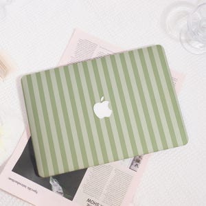 Könnte beinhalten: Eine grün-weiß gestreifte Laptop-Hülle mit dem Apple-Logo in der Mitte. Der Laptop liegt auf einer rosa Zeitung. Das Cover hat ein vertikales Streifenmuster.