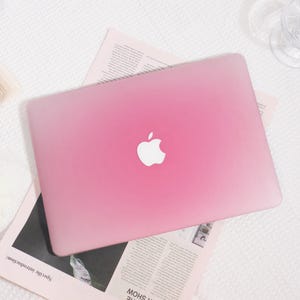 Può includere: Un laptop rosa con il logo Apple bianco è al centro dell'immagine. Il laptop è posizionato sopra un giornale. Il colore del laptop sfuma da un rosa più chiaro nella parte superiore a un rosa più scuro nella parte inferiore.