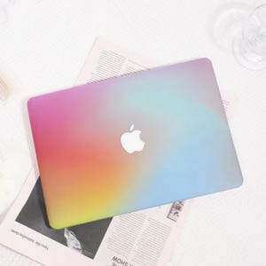 May include: A laptop cover featuring a smooth gradient transition of colours, blending vibrant pink and orange on the left into a soft yellow, transitioning towards a light blue and purple hue on the right, with a central white Apple logo cutout.