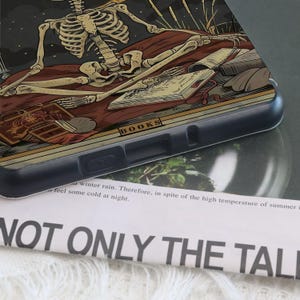K&ouml;nnte beinhalten: Schwarze Handyh&uuml;lle mit einer detaillierten Illustration eines Skeletts, das sich auf einem Bett mit B&uuml;chern ausruht. Das Design enth&auml;lt das Wort "BOOKS" und eine dunkle, gotische &Auml;sthetik. Die H&uuml;lle besteht aus strapazierf&auml;higem Kunststoff.