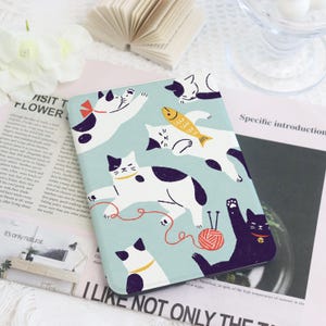 Può includere: Una copertina per quaderno rettangolare azzurra con gatti bianchi e neri. Alcuni gatti tengono in mano un pesce o giocano con il filo. La copertina ha il testo "I LIKE NOT ONLY THE" in basso.