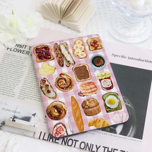 May include: A pink and white checkered notebook featuring watercolor illustrations of various breakfast foods, including toast with jam, croissants, and a cup of coffee. The text "I LIKE NOT ONLY THE" is visible at the bottom.