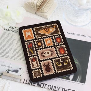 Puede incluir: Estuche rectangular negro con un collage de sellos postales de estilo vintage. Los sellos presentan ilustraciones variadas, incluyendo una polilla, una calavera y flores. El texto "antique dolce 2822" es visible. Disposición en cuadrícula.