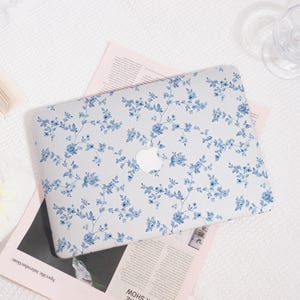 May include: A laptop case with a floral design. The case is light gray with a pattern of blue flowers and green leaves. The Apple logo is visible in the center. The case is on top of a pink magazine.