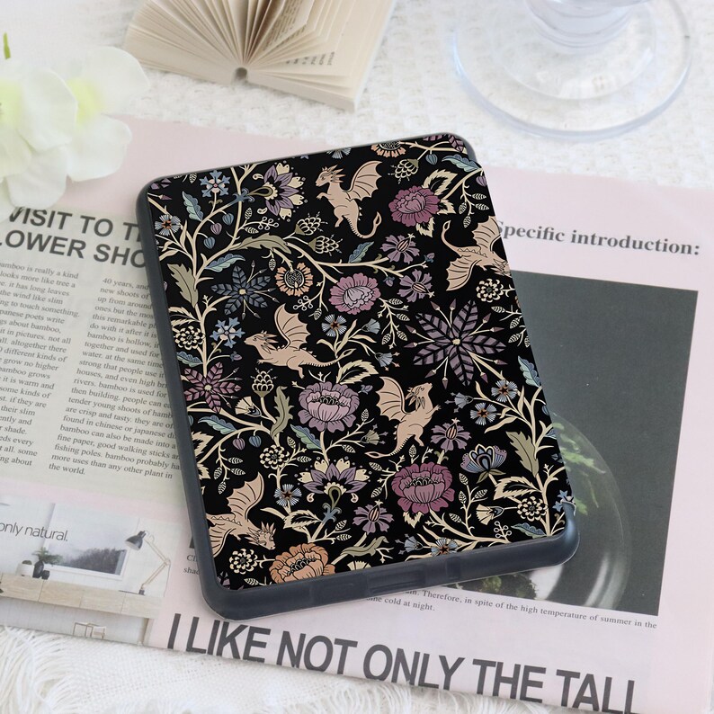 May include: A black phone case featuring a floral and dragon design. The case has a dark background with a pattern of flowers in shades of purple, pink, and beige, along with beige dragons. The case is rectangular.