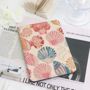 May include: A rectangular, cream-colored case featuring a pattern of colorful seashells in shades of pink, red, blue, and yellow. The case is placed on top of a magazine with text. The overall theme is coastal and beach-inspired.