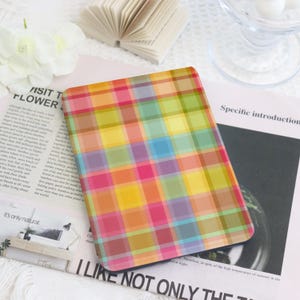 May include: A colorful plaid pattern covers a rectangular object, featuring a vibrant mix of pink, yellow, green, and blue. The object is set on a white surface with a magazine and a small open book in the background. The overall theme is a decorative item.