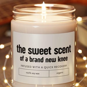 Könnte beinhalten: Eine brennende Kerze in einem klaren Glasgefäß mit einem weißen Etikett, das den schwarzen Text trägt: the sweet scent of a brand new knee, INFUSED WITH A QUICK RECOVERY, 100% soy wax und organic. Die Kerze steht auf einem runden Holzuntersetzer.