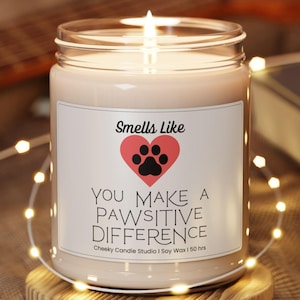 May include: A lit soy wax candle in a clear glass jar with a white label featuring a red heart, a black paw print, and the text: Smells Like YOU MAKE A PAWSITIVE DIFFERENCE Cheeky Candle Studio | Soy Wax | 50 hrs. The candle sits on a round wooden base.