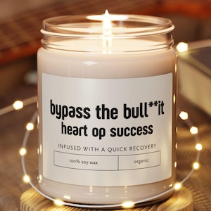 May include: A lit soy wax candle in a clear glass jar with a white label that reads: bypass the bull**it heart op success, infused with a quick recovery, 100% soy wax, organic. The candle sits on a round wooden coaster surrounded by string lights.
