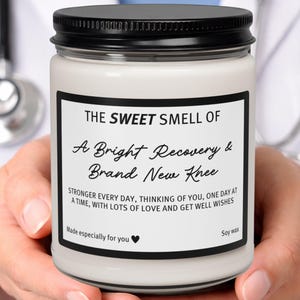 Könnte beinhalten: Eine weiße Sojawachskerze in einem klaren Glasgefäß mit schwarzem Deckel. Das Etikett lautet: THE SWEET SMELL OF A Bright Recovery & Brand New Knee. STRONGER EVERY DAY, THINKING OF YOU, ONE DAY AT A TIME, WITH LOTS OF LOVE AND GET WELL WISHES. Made especially for you.