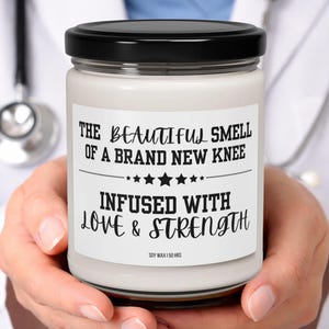 Könnte beinhalten: Ein durchsichtiges Kerzenglas mit schwarzem Deckel und weißem Wachs. Das Etikett trägt den schwarzen Text: THE BEAUTIFUL SMELL OF A BRAND NEW KNEE INFUSED WITH LOVE & STRENGTH, mit einer Sternenreihe und dem Text SOY WAX | 50 HRS darunter.