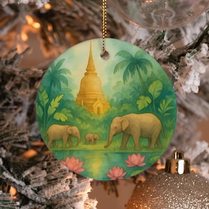 Könnte beinhalten: Ein rundes Keramikornament mit einer gemalten Szene von drei Elefanten, die an einem Teich mit rosa Lotusblüten entlanggehen. Im Hintergrund erhebt sich eine goldene Pagode über üppigen grünen Palmen und Laub unter einem hellen Himmel.