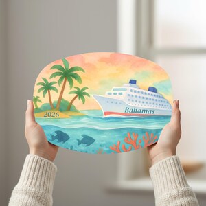 May include: An oval-shaped decorative plaque featuring a watercolour illustration of a cruise ship labelled Bahamas sailing on blue water. The scene includes a tropical island with palm trees, orange coral, and the year 2026 printed on the sandy shore.